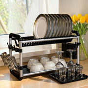 A stainless steel and black plastic dish rack on a kitchen counter, holding stacked plates, bowls, cups, and wine glasses available at spotngrab 